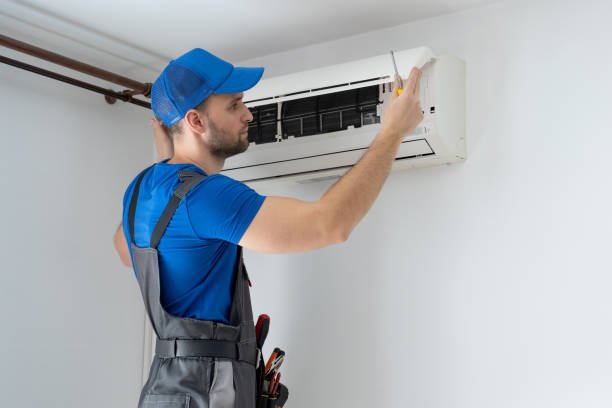 A man wearing overalls and a hat repairs an air conditioner, focused on his task in a residential setting during AC installation in Glendora and Oakland, CA.